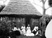 Grupo de quatro mulheres adultas e duas crianças do sexo feminino a bordar, junto a uma casa de madeira com cobertura de colmo, em local não identificado