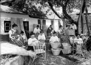 Grupo de mulheres e homens executando trabalhos de artesanato em vime, no quintal de uma casa, em sítio não identificado