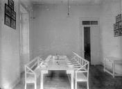 Sala de refeição, vendo-se uma mesa com cadeiras corridas, dezanove pratos sobre a mesa e uma panela ao centro