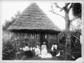 Grupo de quatro mulheres adultas e uma criança do sexo feminino a bordar, junto a uma casa de madeira com cobertura de colmo, em local não identificado